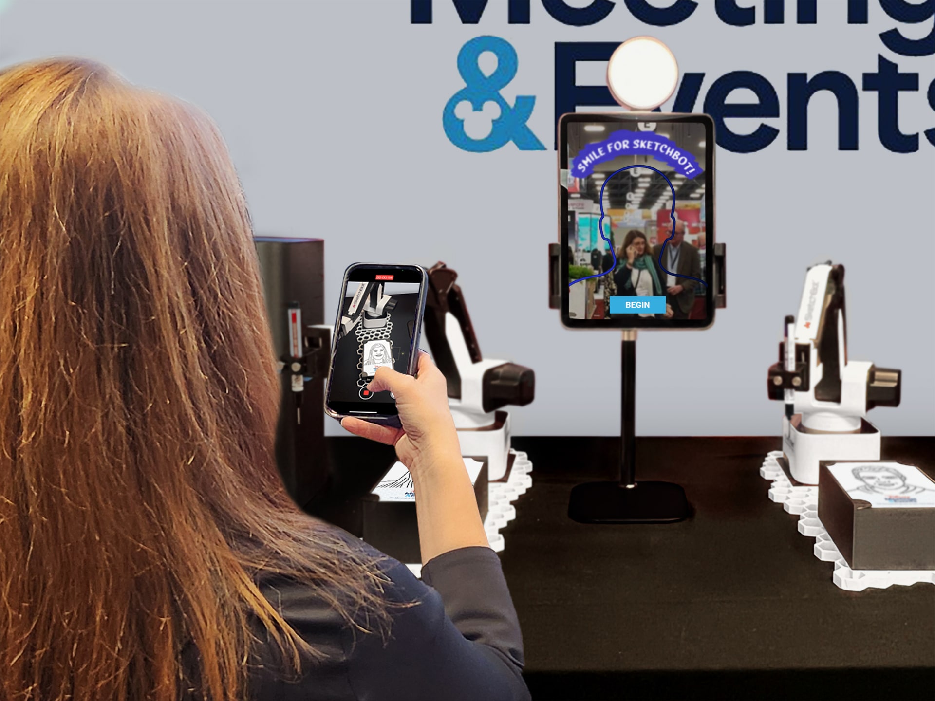 Attendee recording a video as a SketchBot draws their portrait. A selfie kiosk and another SketchBot sit on the table nearby